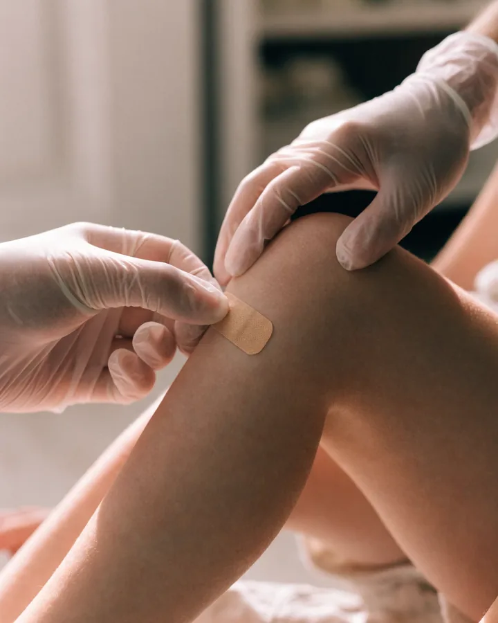 Mãos com luva aplicando um curativo limpo no joelho de uma criança
